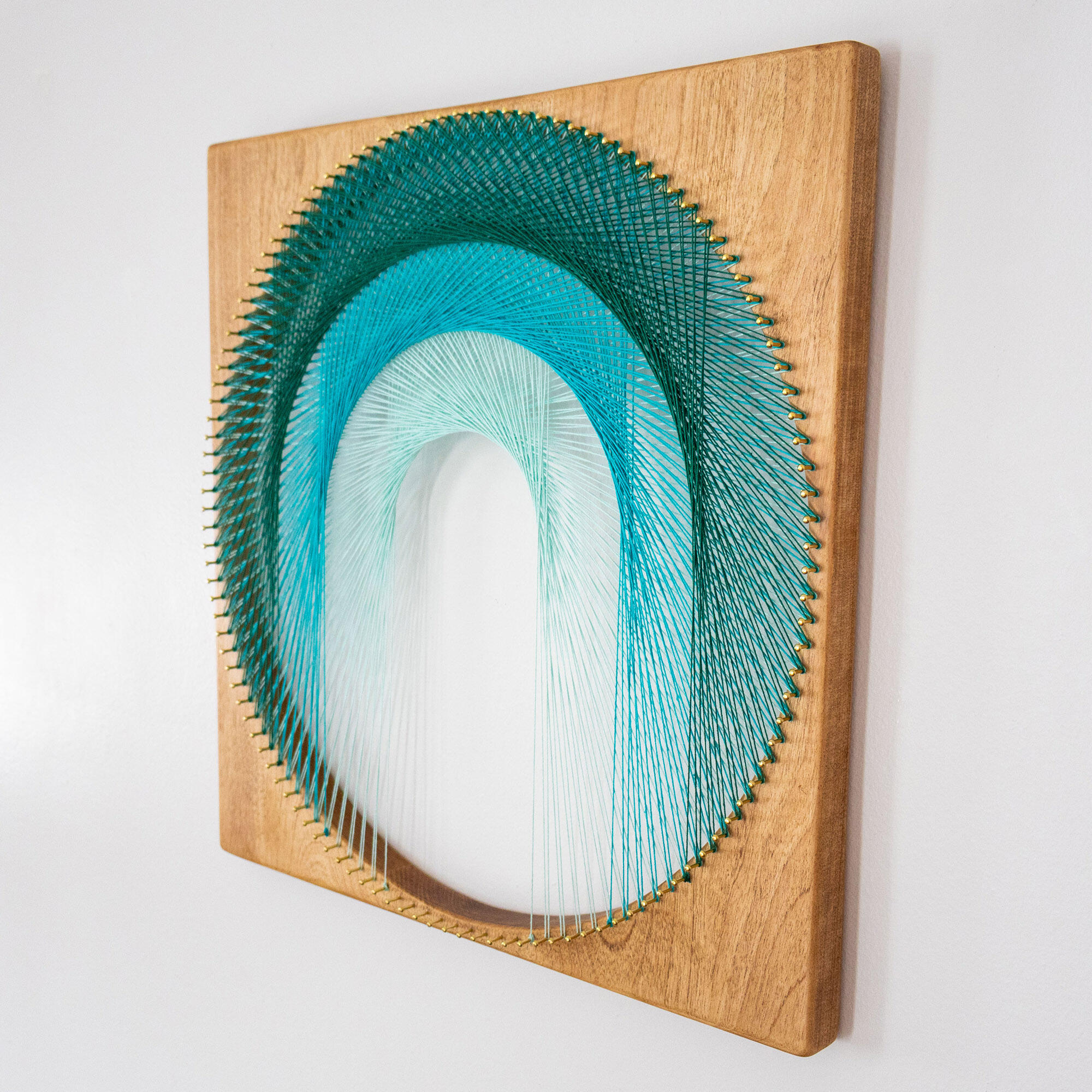 A square birch wood wall art piece featuring a sculptural string‑art arch. Green‑to‑blue cotton threads form a smooth gradient as they wrap around 120 small brass pins, creating depth, shadow, and a refined mid‑century modern look.