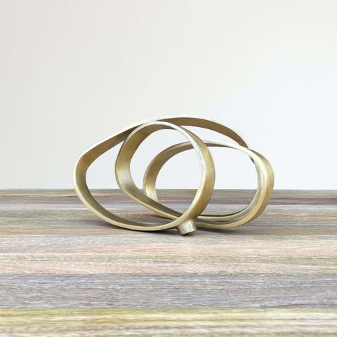  Iron sculpture in gold finish, resembling ribbon intertwined into 3 circles on wood table.