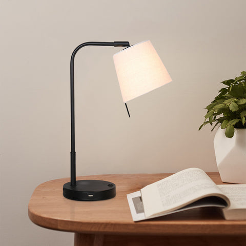 Black arm & off-white linen shade task lamp with a black base including a USB port on a table in a room.