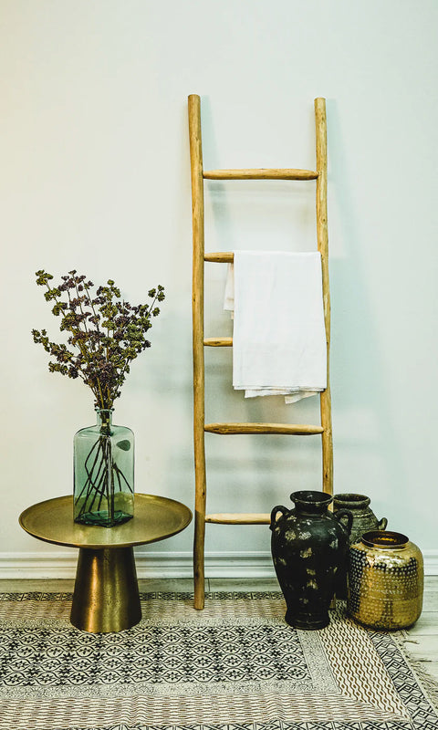 Decorative rustic ladder in natural eucalyptus wood. Towel hanging on one rung, side table with vase on left, & 3 decorative vases on floor on right.