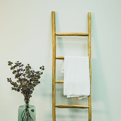 Decorative rustic ladder in natural eucalyptus wood with a towel hanging on one rung and a plant in a glass vase beside it.